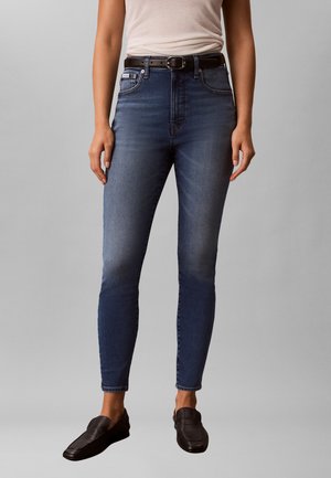 Femme portant un jean skinny bleu foncé, une ceinture noire, un top beige sans manches et des mocassins noirs, debout devant un fond gris.