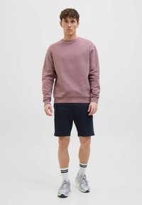 Lyserød sweatshirt med rund hals og ribbede manchetter, parret med marineblå shorts. Modellen har hvide sokker med sorte striber og grå sneakers på.