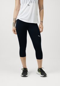 Leggings capri bleu marine avec logo blanc, associés à un haut blanc ample et des chaussures de sport noires avec des accents néon.