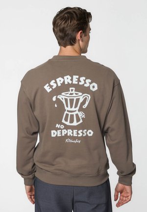 Mand iført brun sweatshirt med hvid kaffekande grafik og tekst "Espresso No Depresso" på bagsiden, stående mod en ensfarvet baggrund.
