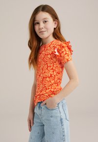 Apelsīnu krāsas ziedu raksta T-krekls ar īsām volānu piedurknēm, apvienots ar gaiši ziliem džinsiem, kuriem ir izšūtas margrietiņas.