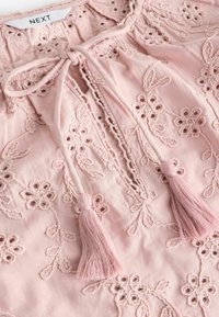 Blusa rosa chiaro con motivi floreali ricamati, scollatura con lacci e due nappine rosa alle estremità. Tessuto leggero.