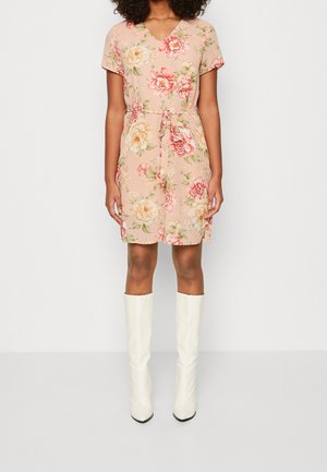 Kobieta ubrana w beżową, kwiatową sukienkę do kolan z krótkimi rękawami i przewiązanym paskiem w talii, zestawioną z białymi kozakami za kolano.