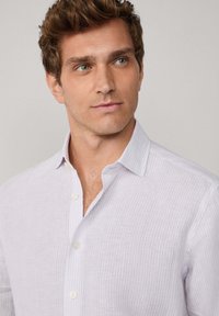 Junger Mann mit kurzen braunen Haaren, der ein helllila gestreiftes Button-Up-Hemd trägt und leicht nach rechts schaut, vor einem schlichten Hintergrund.