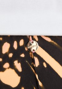 Schwarz-orange gemusteter Stoff mit einem metallischen Logodetail. Glatte Textur mit passgenauer Form. Weißer Abschnitt oben.