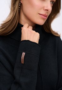 Kobieta w czarnym swetrze z ribbowanymi rękawami z metką "blue shadow", trzymająca kołnierz blisko podbródka, widoczna złota kolczyka w kształcie koła.