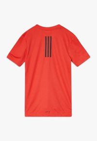 Světlé červené sportovní tričko s krátkým rukávem a třemi svislými černými pruhy na zadní straně, s malým textem "AEROREADY" blízko lemu.