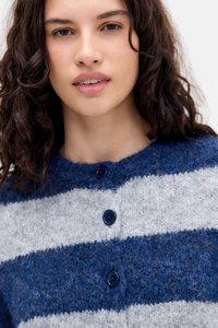Jonge vrouw met krullend bruin haar die een fuzzy blauw- en grijsgestreept knooptrui draagt tegen een effen achtergrond.