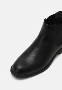 Bottine noire en cuir à enfiler avec un bout arrondi et des panneaux latéraux élastiques sur fond blanc.