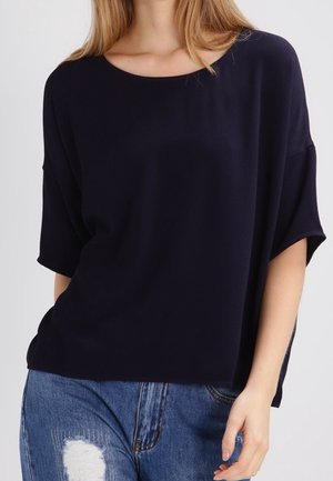Vrouw draagt een loszittend zwart T-shirt met korte mouwen en versleten blauwe spijkerbroek, poseert met één hand bij de zak tegen een effen achtergrond.