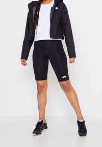 Zwarte waterdichte jas over een wit T-shirt, gecombineerd met zwarte fietsshorts en zwarte sportchoenen. Bevat logo-accenten en heeft een aangesloten ontwerp.