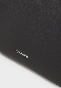 Svart teksturert lærpose med en glatt finish, med en sølvfarget Calvin Klein-logo, rektangulær form og minimalistisk design.