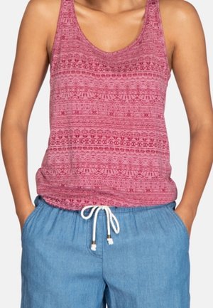 Kvinde iført ærmeløs rød mønstret tanktop stoppet ned i blå snørrebukser med hænderne i lommerne.