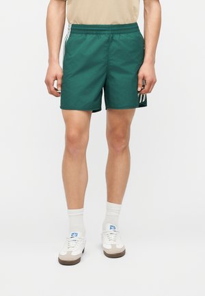 Uomo che indossa pantaloncini sportivi verde scuro con strisce bianche, maglietta beige, calzini bianchi e sneaker Adidas bianche su sfondo neutro.