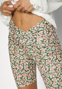 Calças de ciclismo com padrão floral em verde, apresentando flores rosas e brancas, com um design ajustado e tecido texturizado. Destacadas com franzidos laterais.