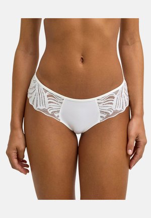 Kobieta ubrana w białe bikini z koronki, z przezroczystymi, wzorzystymi detalami koronki po bokach oraz gładkim, materiałowym panelem na środku.