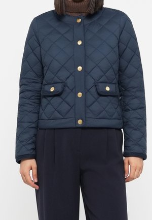 Femme portant une veste matelassée bleu marine avec des boutons dorés et des poches à rabat, par-dessus un pull à col roulé marron et un pantalon foncé.