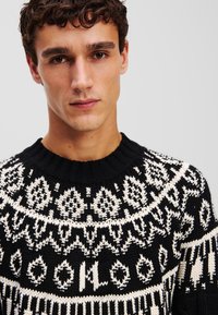 Pull en laine noir et blanc avec un col rond et des motifs géométriques, utilisant des fils texturés et des designs contrastés.