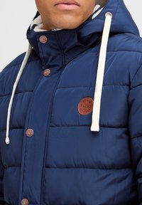 Mørkeblå pufferjakke med et tykt, kviltet design, der har en faux fur-foring, brun logo-lap og beige snørebånd i hætten.