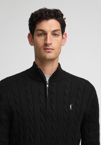 Czarny sweter o splocie warkoczowym z wysokim kołnierzem i zapięciem na zamek, z małym różowym logotypem na piersi. Gładka tekstura.