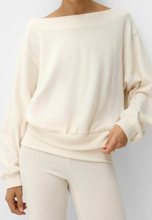 Kobieta ubrana w kremowy sweter z odkrytymi ramionami i długimi rękawami z prążkowanymi mankietami oraz pasem, w komplecie z dopasowanymi kremowymi spodniami.