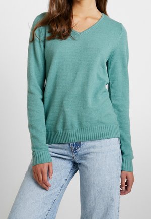 Pull à col en V, à manches longues, de couleur vert aqua, fabriqué en tricot doux. Présente des poignets et un ourlet côtelés. Associé à un jean en denim clair.