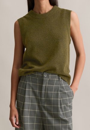 Pull sans manches vert olive associé à un pantalon taille haute gris à carreaux, une main dans la poche, fond neutre.
