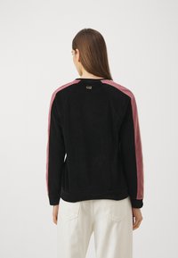 Donna con lunghi capelli castani girata di spalle, che indossa una felpa nera con strisce rosa sulle maniche e pantaloni bianchi, su uno sfondo neutro.
