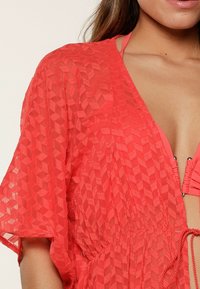 Cubierta ligera de color coral, hecha de tela texturizada, que presenta un escote en V, mangas cortas y una cinturilla ajustable con lazo, con detalles de patrón geométrico.