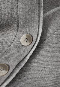 Szürke szövet finom textúrával, sima szegéllyel. A varrás mentén három nagy, világos színű gomb látható.