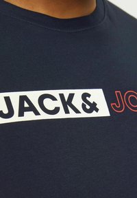 Tamsiai mėlynos spalvos medvilninė marškinėlių suapvalintu kaklu, su balta stačiakampio formos grafika, kurioje užrašyta "JACK&" ryškiai juodai, o "JO" raudonai. Lygi tekstūra.