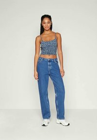 Mulher com tranças longas, vestindo um top crop floreado azul, calças jeans largas de cintura alta azuis e ténis brancos, em pé sobre um fundo simples.