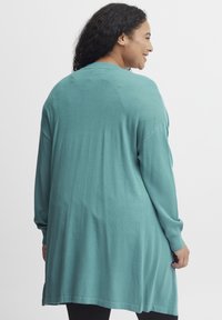 Långärmad teal cardigan med en avslappnad passform, mjuk struktur, medellång längd, sidofickor och ribbade ärmslut. Visad bakifrån.