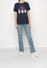 Kvinna iförd en marinblå jult-shirt med tre pingviner och texten "God Jul", tillsammans med ljusblå jeans och vita sneakers.