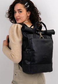 Nainen, jolla on kihara hius, pukeutunut beigeen neuleeseen ja bežezapariin, kantaa mustaa, rullakorkkista nahkareppua, jossa on kultainen solki ja "Expatrié"-logo.