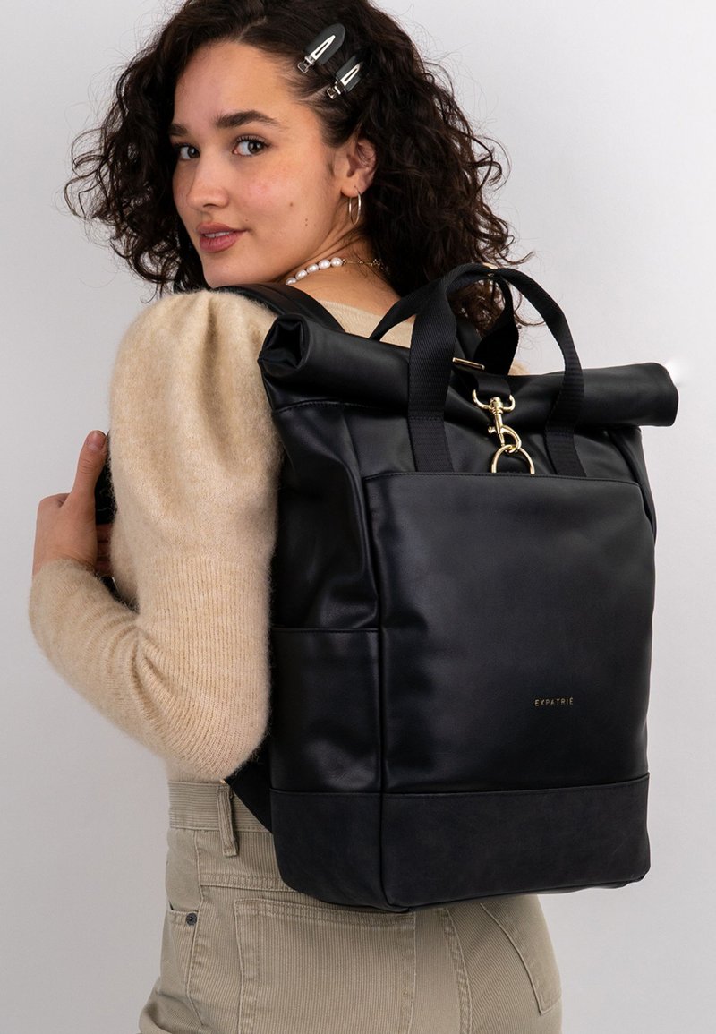 Nainen, jolla on kihara hius, pukeutunut beigeen neuleeseen ja bežezapariin, kantaa mustaa, rullakorkkista nahkareppua, jossa on kultainen solki ja "Expatrié"-logo.