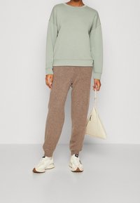 Sudadera verde claro con hombros caídos, combinada con pantalones jogger marrones y zapatillas blancas. Sosteniendo un bolso triangular color crema.