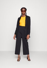 Blazer noir sur un haut orange vif, associé à un pantalon noir large et des sandales à talons noires. De grandes lunettes de soleil brunes complètent le look.