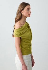 Haut asymétrique vert en tissu doux, avec une texture drapée sur une épaule, associé à un pantalon blanc ajusté. Des boucles d'oreilles en or rehaussent le look.