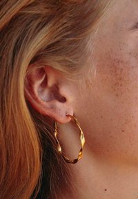 Goldene, gedrehte Creole mit wellenförmigem Design. Die Ohrringe werden durch einen kleinen Stift befestigt und haben eine polierte metallische Oberfläche.