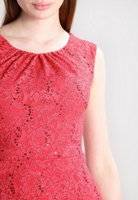 Robe en dentelle corail vif avec manches cape, dotée d'un col froncé et d'accents de sequins pour plus de texture et de détails.