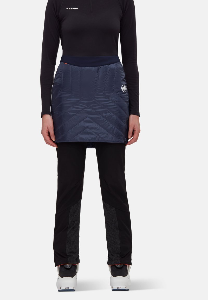 Isolerad, marinblå vadderad skort med en slät textur och elastisk midja. Bärs över svarta byxor, har logodetalj på sidan.