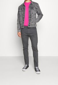 Szara kurtka dżinsowa na różowej koszulce, w zestawie z szarymi, wąskimi spodniami oraz czarnymi trampkami z płótna z białymi podeszwami. Jednolite tło.