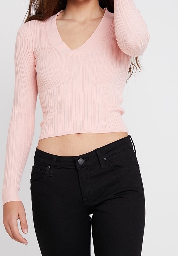 Růžový žebrovaný svetr s hlubokým výstřihem do V a dlouhými rukávy, kombinovaný s vysokým pasem černých džín, který zdůrazňuje přiléhavý střed těla.