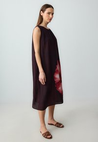 Ärmelloses dunkelrot Kleid mit einem großen roten Blumenmuster auf einer Seite, aus leichtem Stoff gefertigt und mit lockerer Passform. Braune Rutsch-Sandalen.