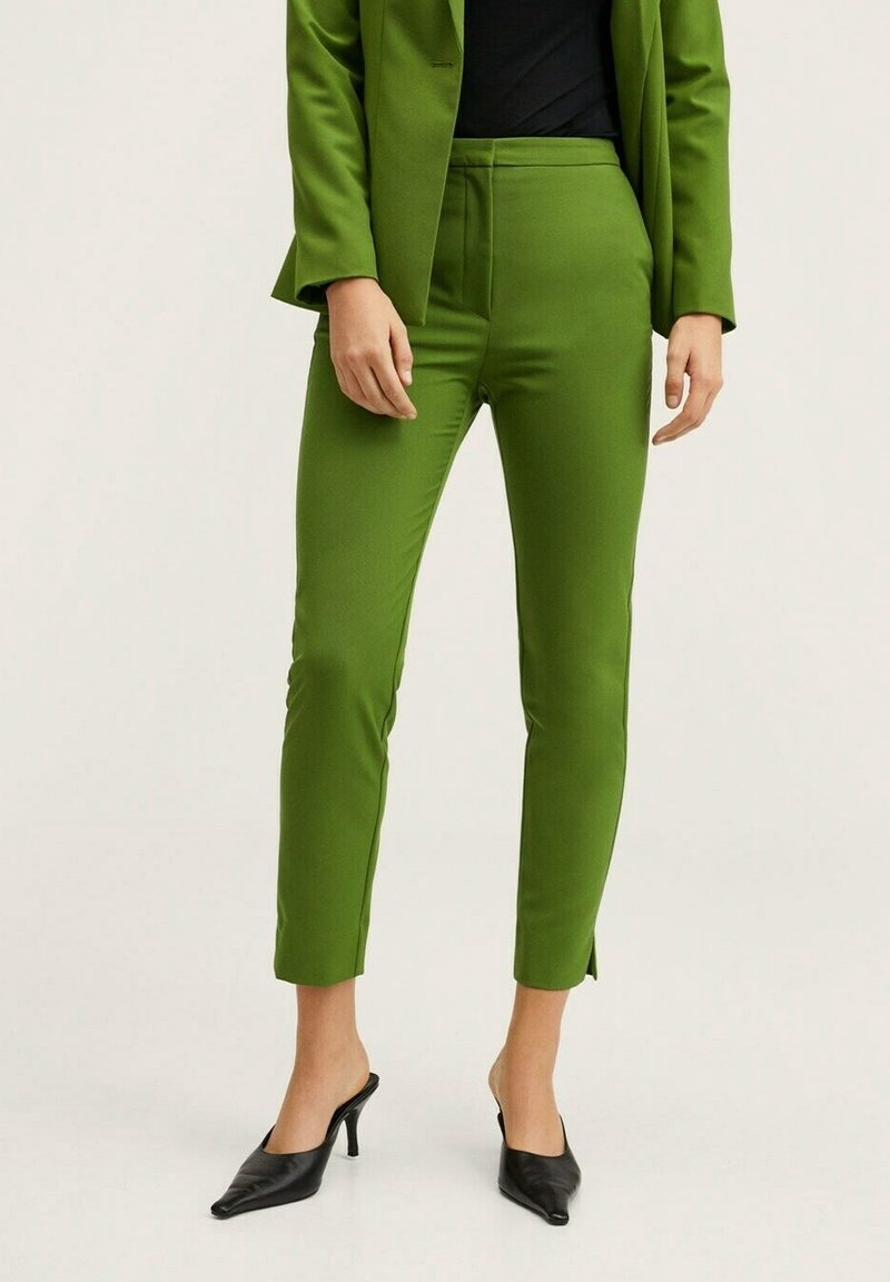 Personne portant un pantalon olive ajusté avec des fentes aux chevilles, un blazer assorti, un haut noir et des mules à talons noires à bout pointu.
