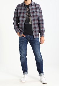 Camisa de franela en gris con un patrón de cuadros en rojo y negro, superpuesta a una camiseta gráfica negra. Combinada con jeans de mezclilla azul y zapatillas blancas.
