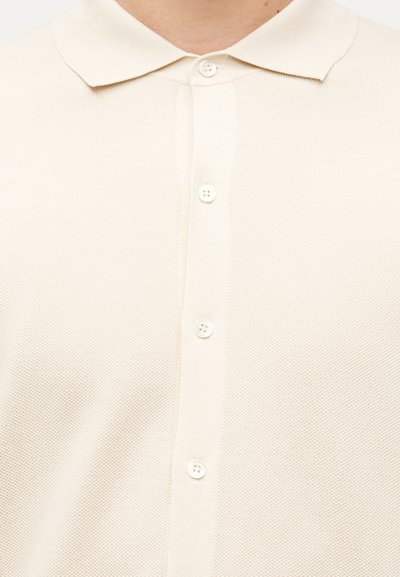 Gros plan d'une personne portant un polo beige texturé avec un col et quatre boutons blancs sur le devant.