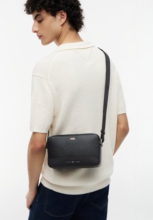 Giovane uomo che indossa una maglia a maniche corte color crema e jeans scuri, con una borsa a tracolla nera Tommy Hilfiger sulla schiena.