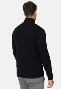 Pull en maille noire avec un col montant, des poignets et un ourlet côtelés. Le tissu semble texturé, adapté aux temps frais.